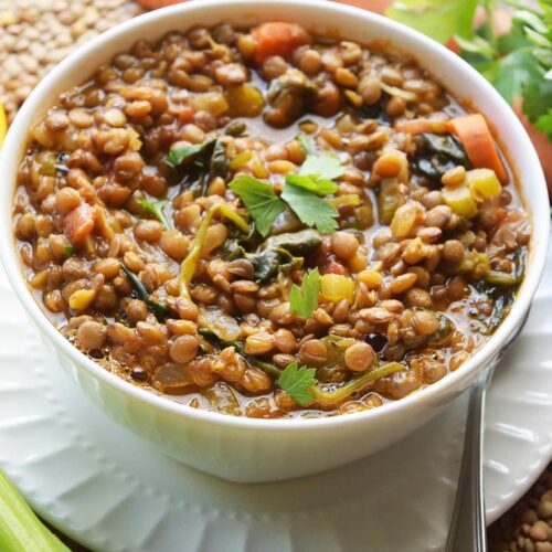 A white bowl filled with Spinach Lentil Soup sits on a saucer plate surrounded by carrots, celery and dried lentils.