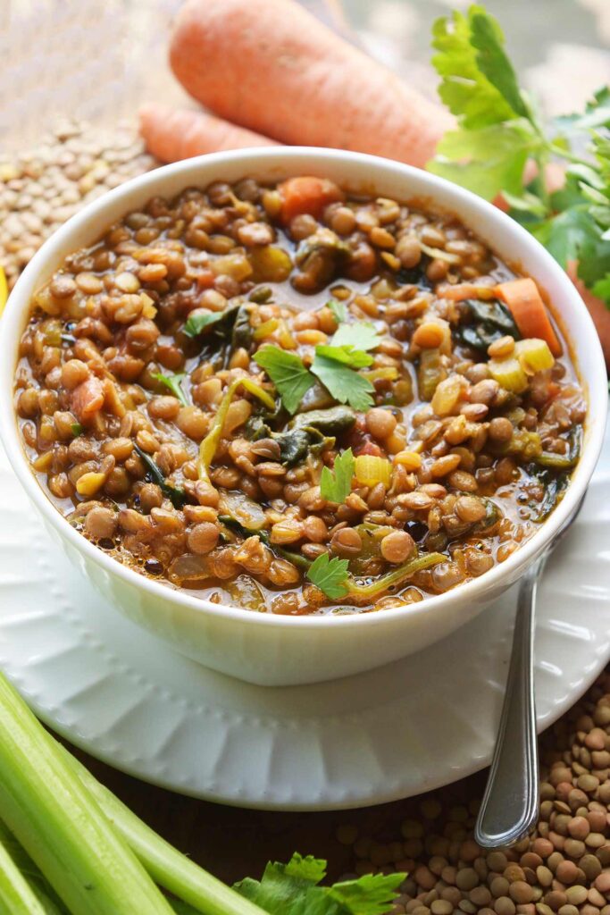 A white bowl filled with Spinach Lentil Soup sits on a saucer plate surrounded by carrots, celery and dried lentils.