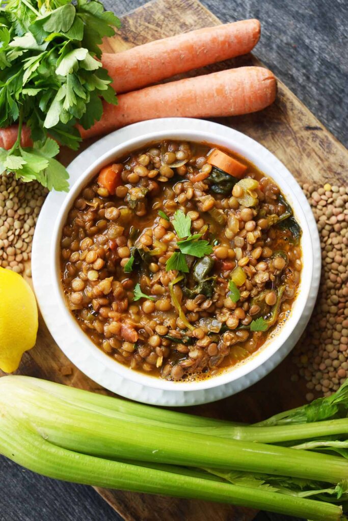 An overhead view of a white bowl filled with Spinach Lentil Soup surrounded by carrots, celery, dried lentils and fresh parsley.