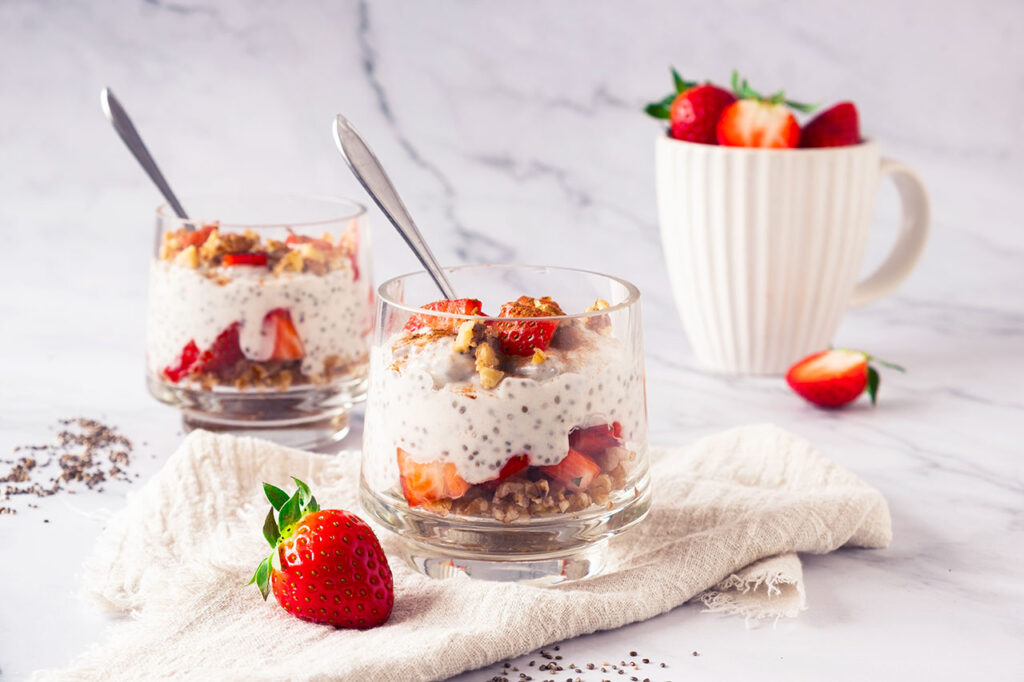 Two clear glasses filled with strawberry chia pudding sitting on a counter with a towel and some spoons.