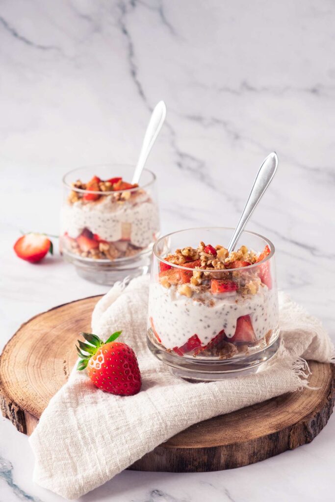 Two glasses on a marble countertop. On on the counter and one on a cutting board. Both are filled with Strawberry Chia Pudding.