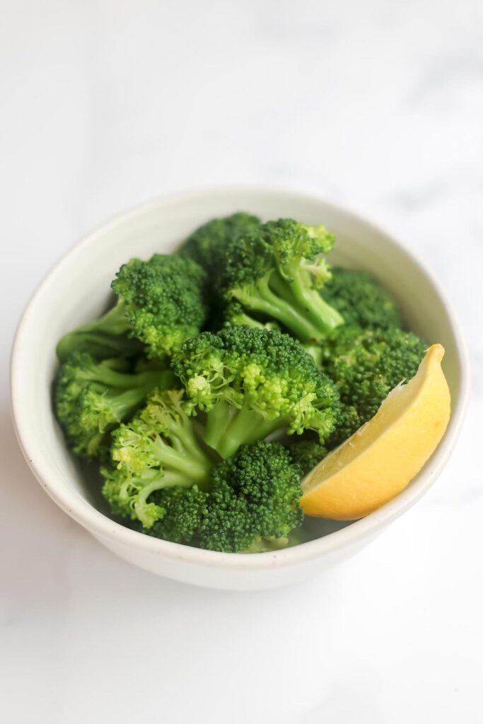 A side view of a white bowl of broccoli with a lemon wedge.