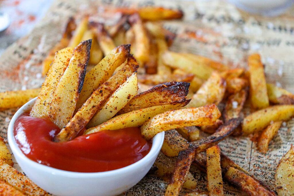 Air Fryer French Fries stuffed into a white bowl of ketchup on a piece of newpaper.