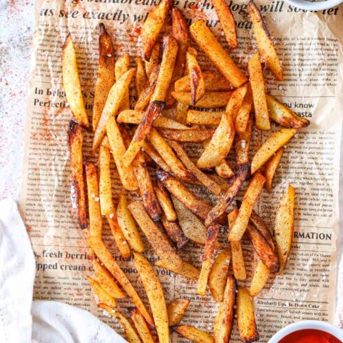 Air Fryer French Fries laying on a piece of newspaper with a bowl of ketchup, a bowl of pepper and a small container of salt.