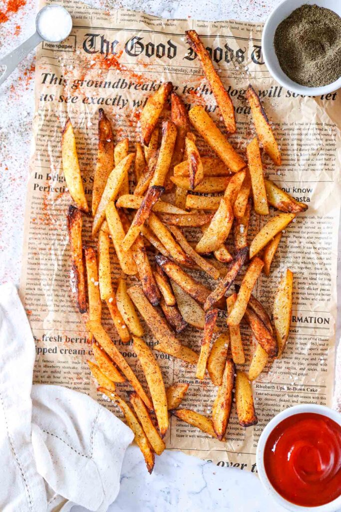 Air Fryer French Fries laying on a piece of newspaper with a bowl of ketchup, a bowl of pepper and a small container of salt.