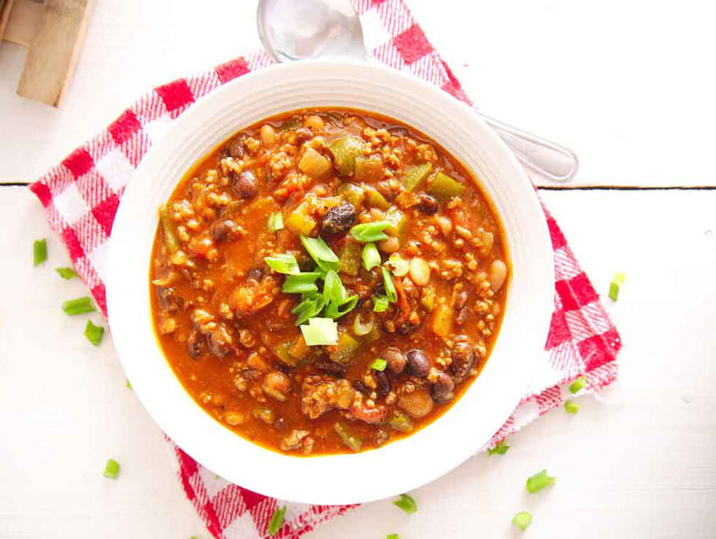 An overhead view of a white bowl filled with beef chili sitting on a red and white checkered cloth on a white, wood table.