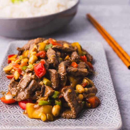 Kung Pao Beef on a platter with chopsticks and a bowl of rice in the background.