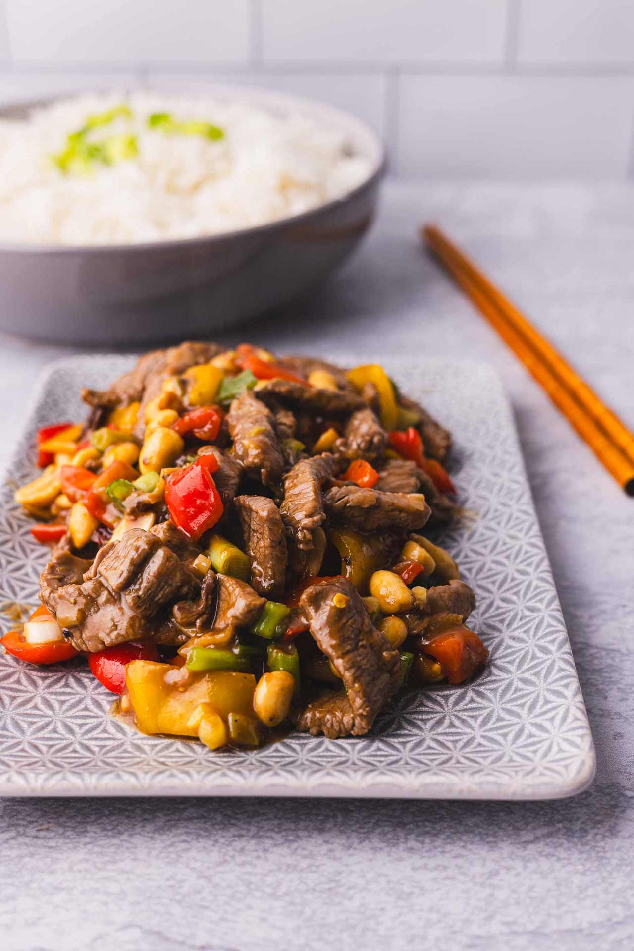 Kung Pao Beef on a platter with chopsticks and a bowl of rice in the background.