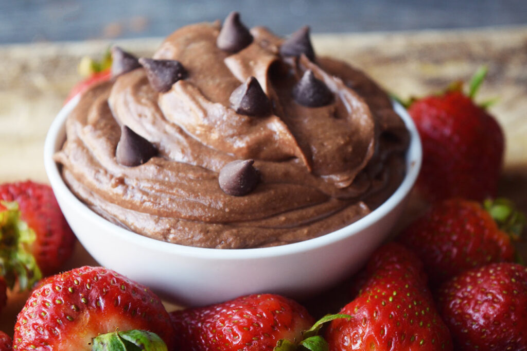 A side view of brownie batter dip in a white bowl surrounded by fresh strawberries.