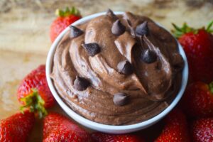 A side view of a white bowl filled with brownie batter dip and surrounded by fresh strawberries.