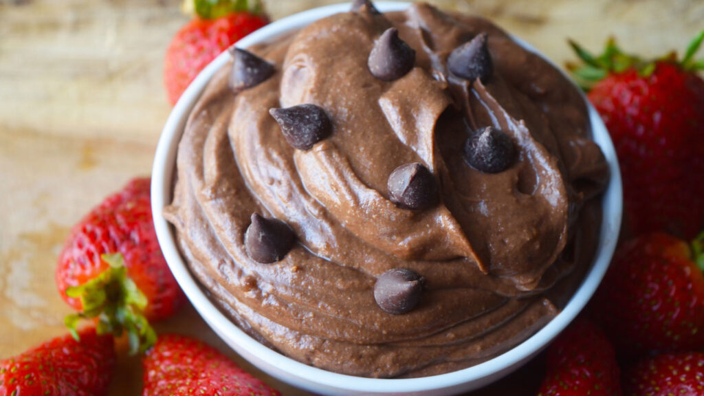A close up view of a white bowl filled with brownie batter dip and surrounded by fresh strawberries.