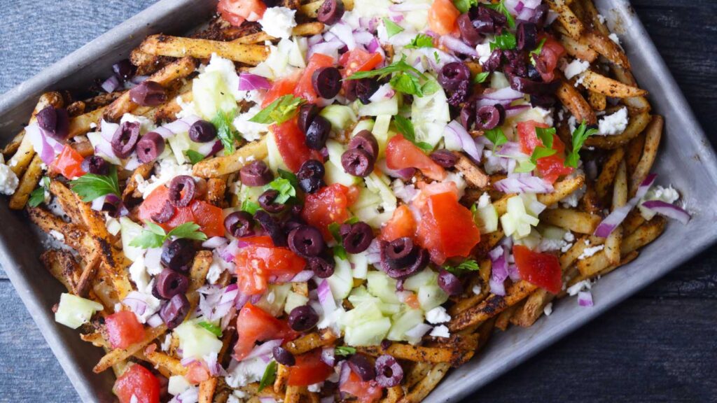 An overhead view of a sheet pan filled with Greek fries and loaded with fresh toppings.