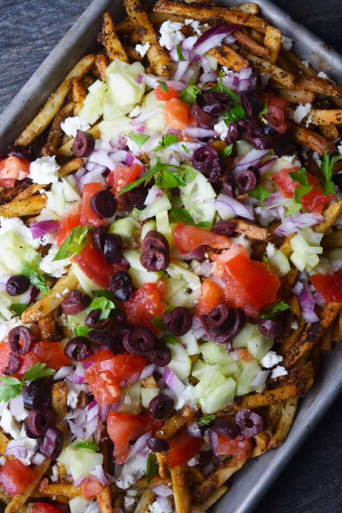 An overhead view of a sheet pan filled with Greek fries loaded with Mediterranean toppings.