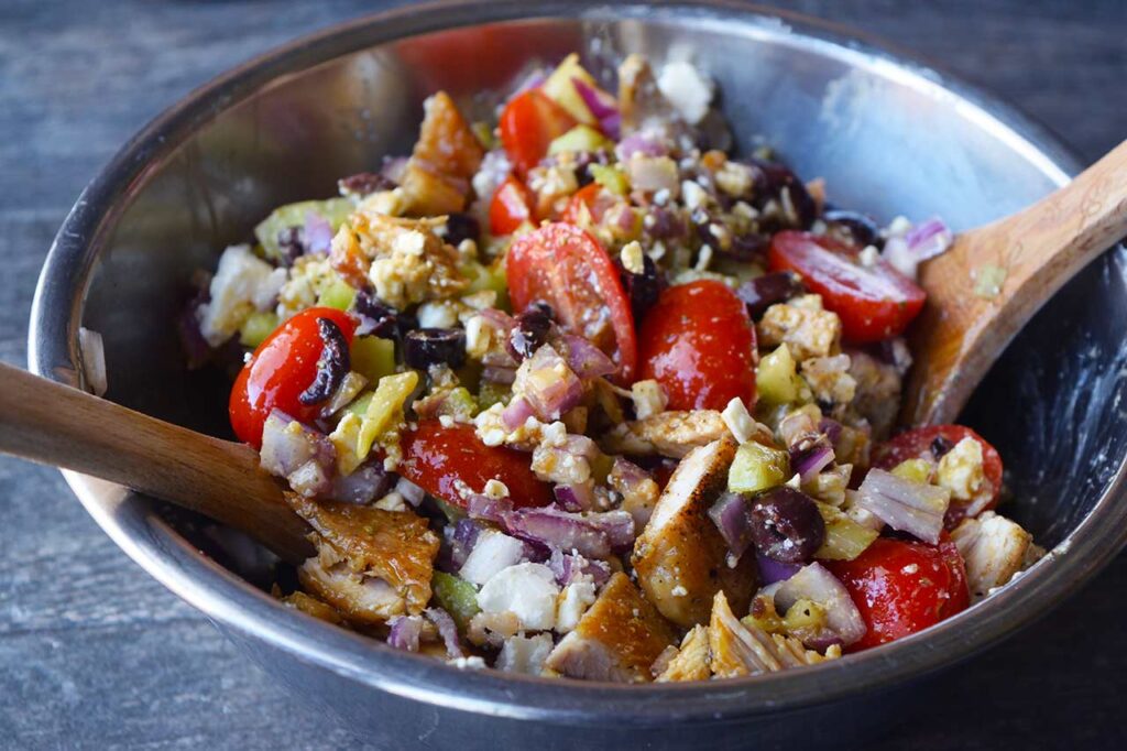 An side view of the mixed Greek chicken salad in a mixing bowl with wood salad tongs.