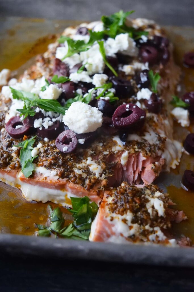 A front view of a salmon fillet on a baking sheet topped with olives, feta cheese and fresh parsley.