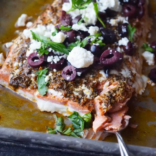 A salmon fillet on a baking sheet topped with olives, feta and fresh parsley. A fork holds a small piece of it and rests in front of the fillet.