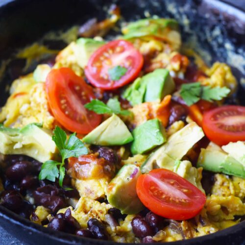 A black, cast iron skillet filled with Mexican Scrambled Eggs garnished with tomatoes, avocado, salsa, and fresh herbs.