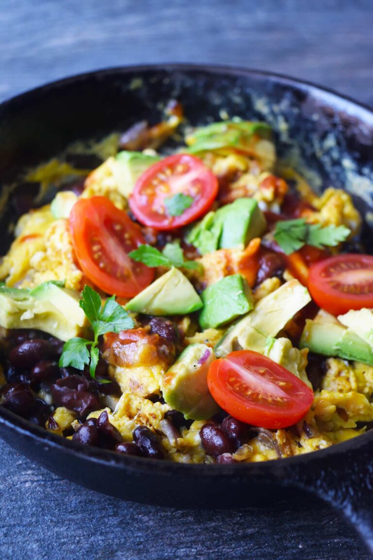 A black, cast iron skillet filled with Mexican Scrambled Eggs garnished with tomatoes, avocado, salsa, and fresh herbs.