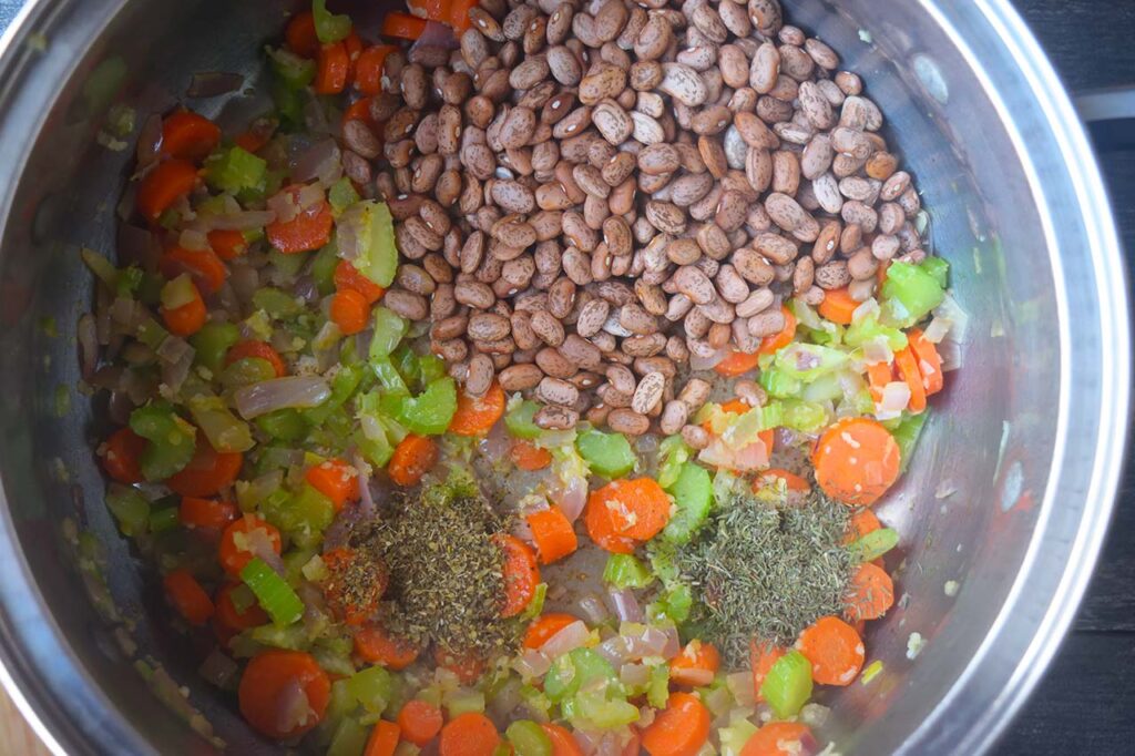 Spices and dry pinto beans added to cooked veggies in a large soup pot.