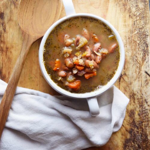 A write crock of Pinto Bean Soup sits on a cutting board with a wooden spoon and a white towel.