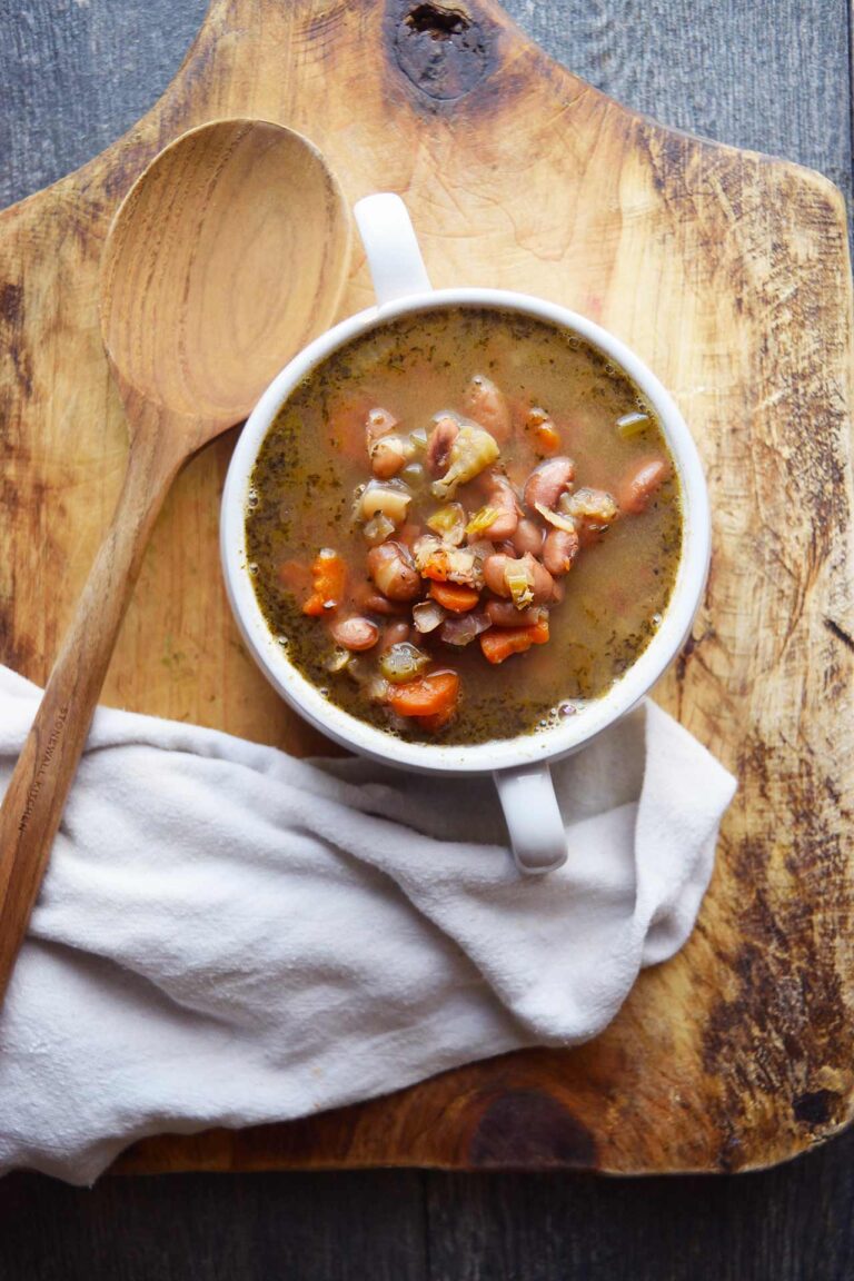 A write crock of Pinto Bean Soup sits on a cutting board with a wooden spoon and a white towel.