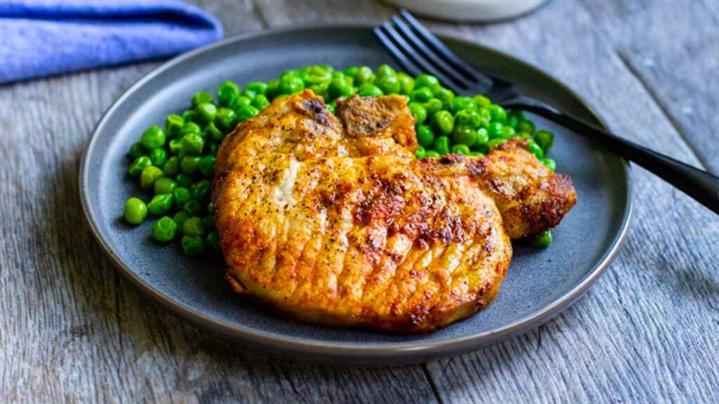 A plate of green peas and a golden brown pork chop.
