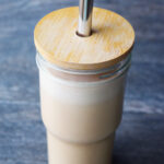A side view of a glass with wood lid and metal straw filled with Almond Butter Almond Milk.