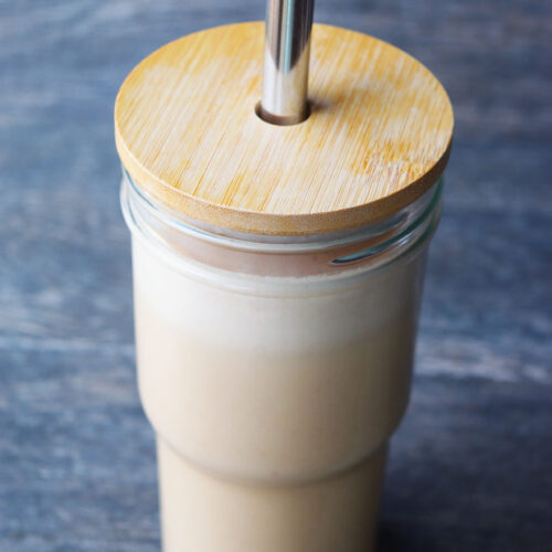 A side view of a glass with wood lid and metal straw filled with Almond Butter Almond Milk.