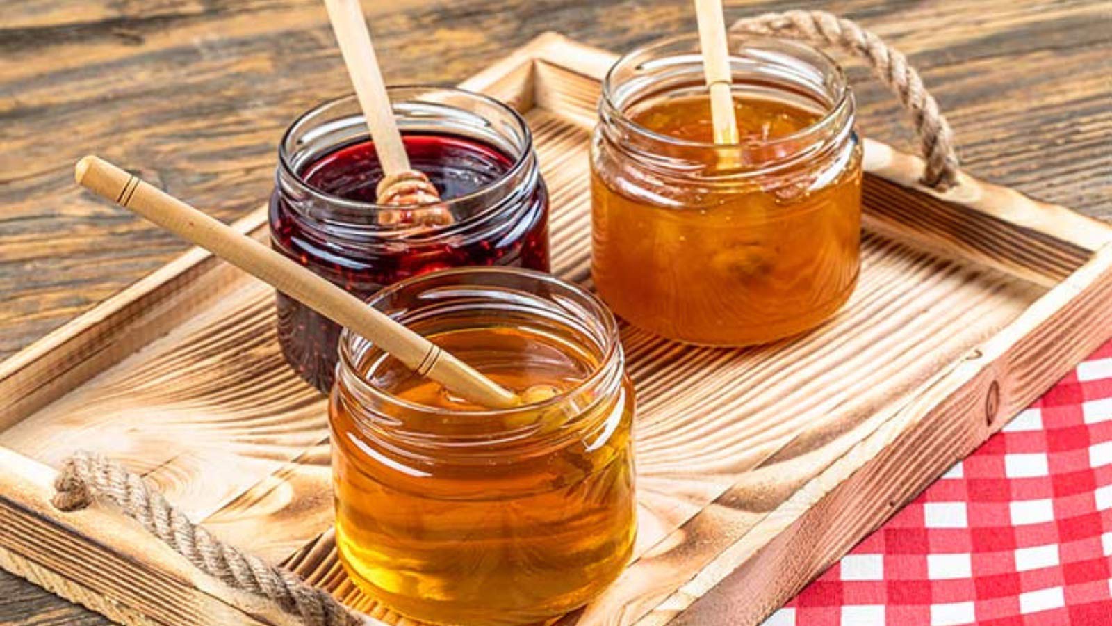Three jars containing jams and honey on a wood serving tray.