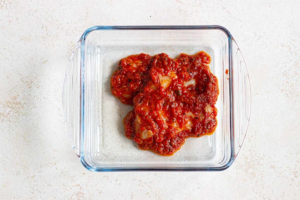 A chicken breast smothered in blended roasted red peppers in a casserole dish.