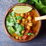 An overhead view of a wood bowl filled with Chickpea Curry and garnished with fresh, chopped cilantro and a lime wedge.