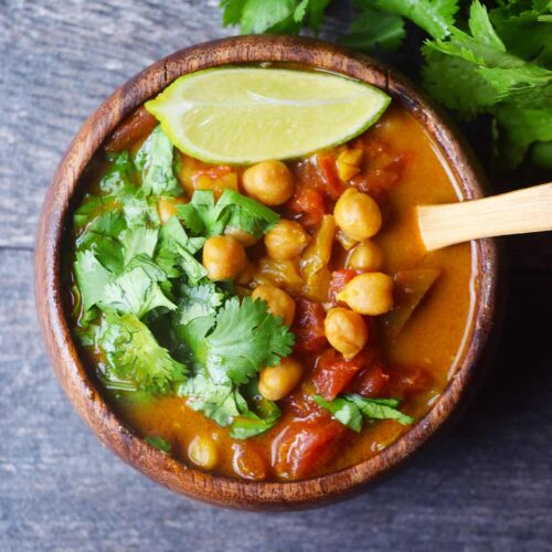 An overhead view of a wood bowl filled with Chickpea Curry and garnished with fresh, chopped cilantro and a lime wedge.