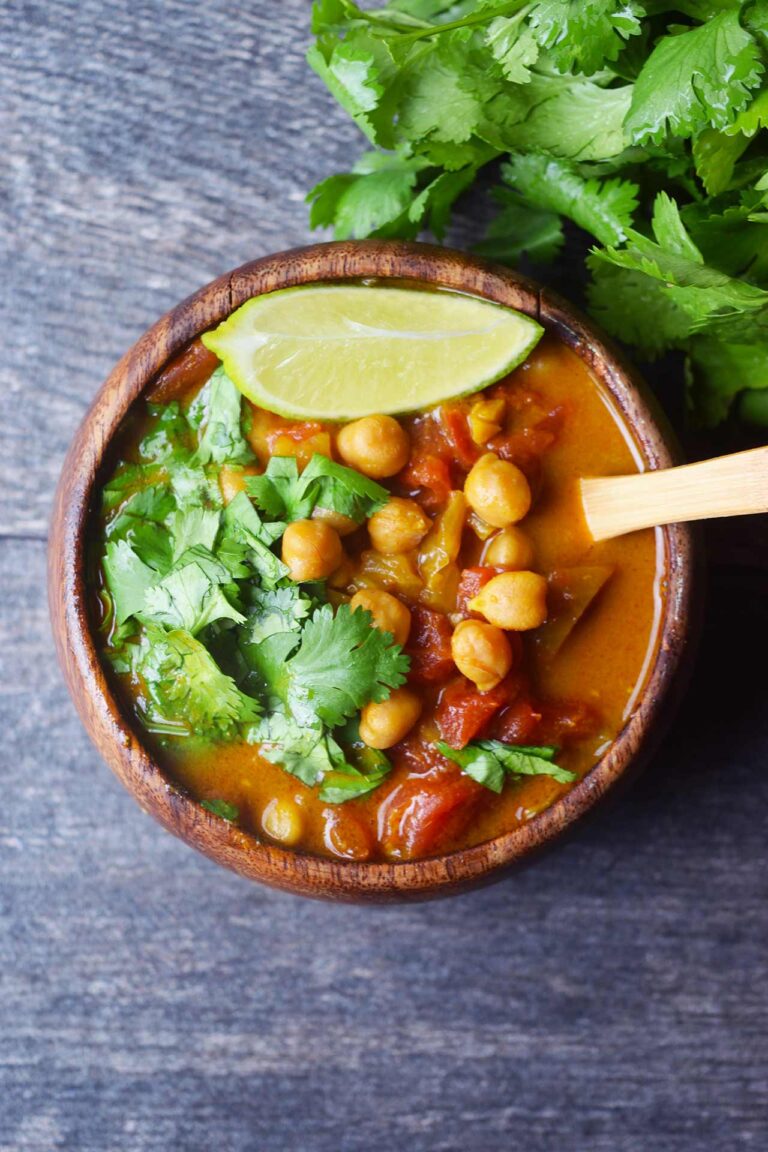 An overhead view of a wood bowl filled with Chickpea Curry and garnished with fresh, chopped cilantro and a lime wedge.