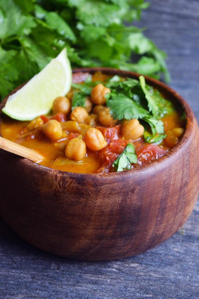 A side view of a wood bowl filled with Chickpea Curry. A bunch of fresh cilantro lays behind the bowl.
