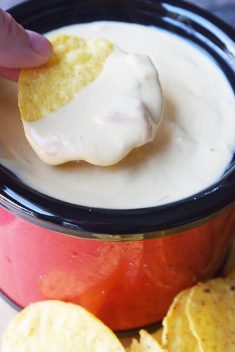A round corn chip dipped in vegan queso from a red and black crock.
