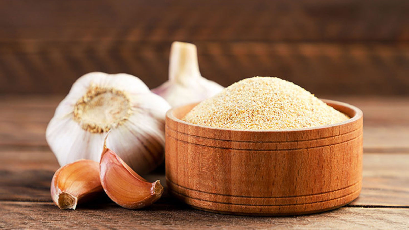A wood bowl filled with garlic powder. A head of garlic and some cloves lay next to it.