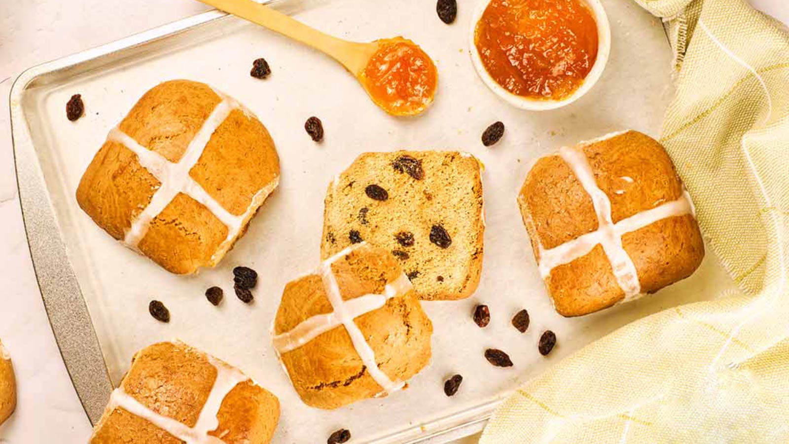 An overhead view of hot cross buns on a baking pan with raisins strewn around them and a bowl of jam sits to the side.