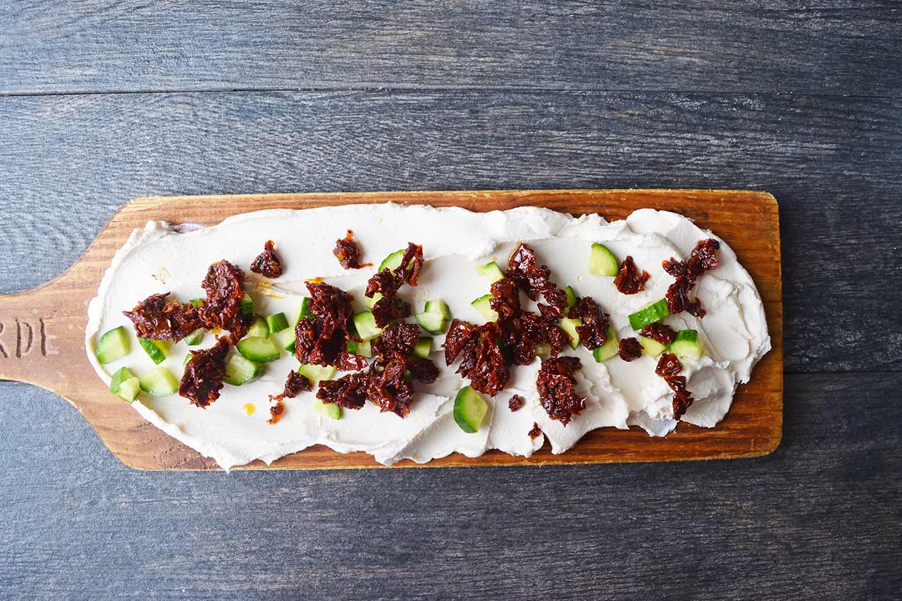 An overhead view of a charcuterie board with chopped cucumbers and chopped sun-dried tomatoes sprinkled over the cream cheese.