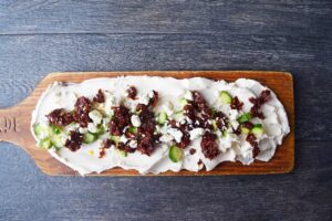 Feta cheese crumbles sprinkled over chopped cucumber and sun-dried tomatoes over cream cheese on a charcuterie board.