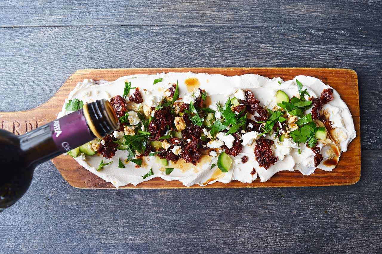 Garnishing a Mediterranean Cream Cheese Dip board with a sprinkle of balsamic vinegar.