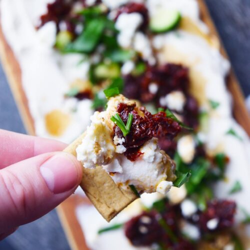 A hand holds a cracker with a scoop of Mediterranean Cream Cheese Dip on it.