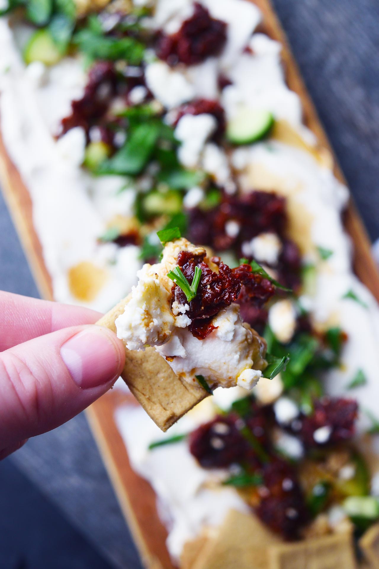 A hand holds a cracker with a scoop of Mediterranean Cream Cheese Dip on it.