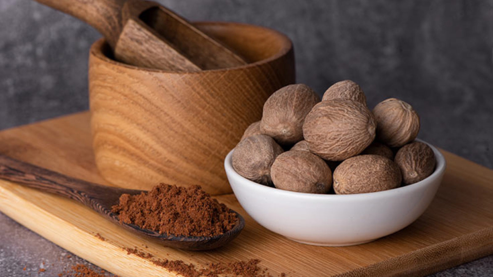 A bowl of nutmegs sits next to a spoon full of ground nutmeg.