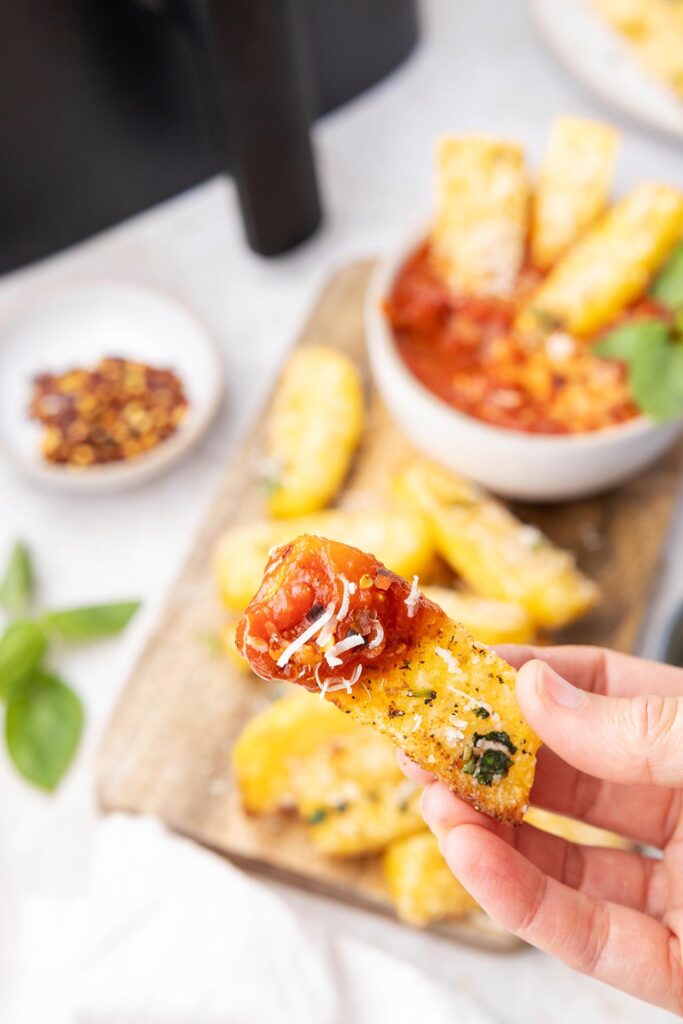 A hand holds a polenta fry that has been dipped in marinara sauce.