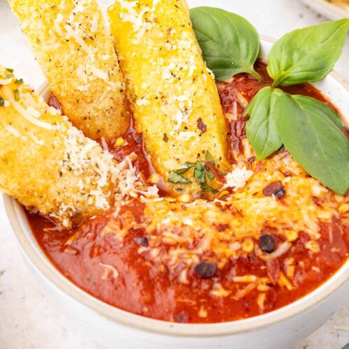 Three polenta fries sitting in a bowl filled with marinara sauce.