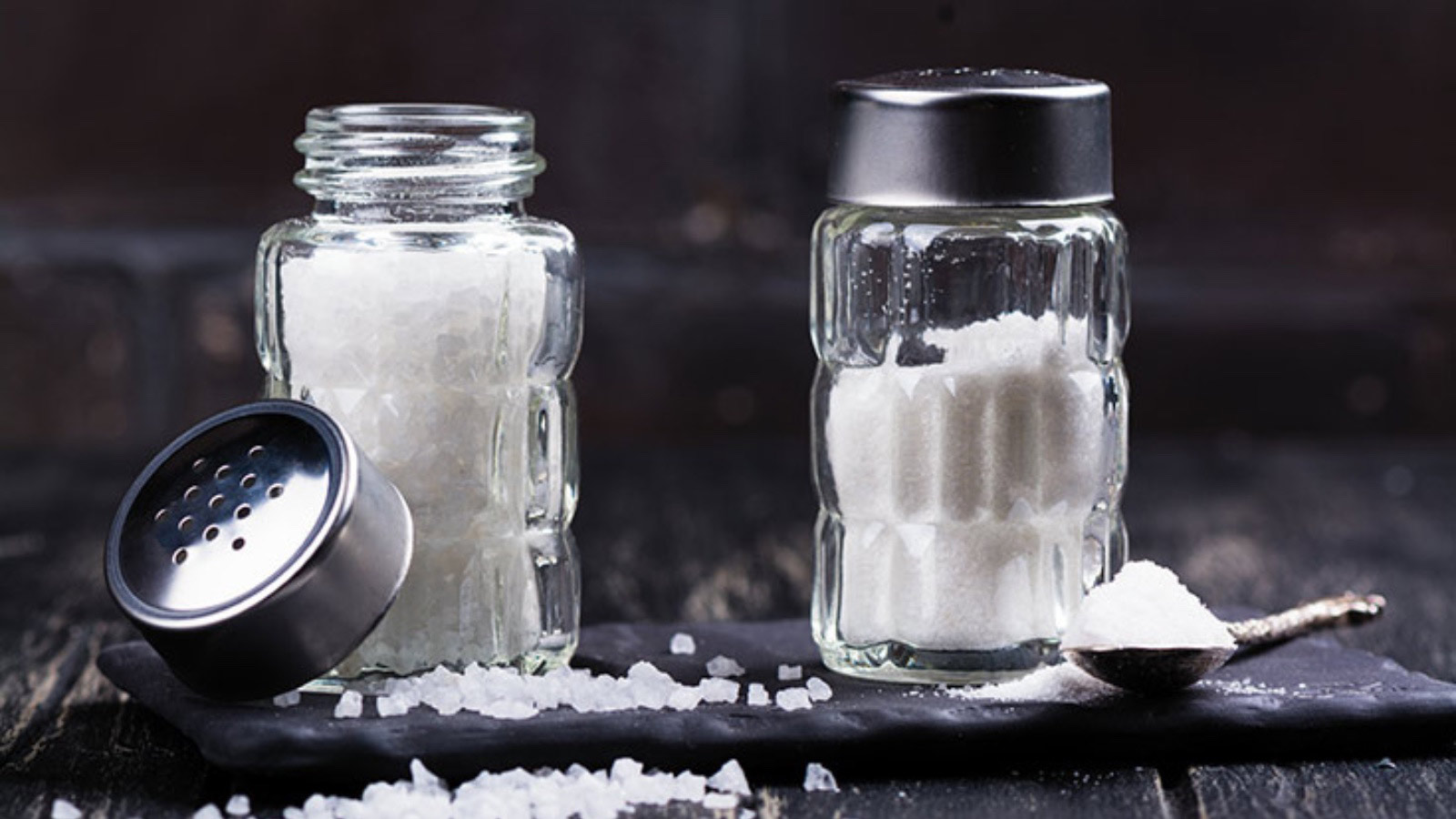 Glass salt shakers with sea salt coarse and fine on dark wooden table.