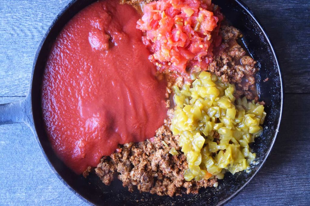 Tomatoes, tomato sauce and green chilies added to seasoned, ground meat in a skillet.