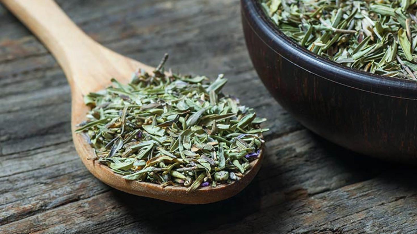 A bowl and wooden spoon sit on a wooden table and are filled with dried thyme leaves.