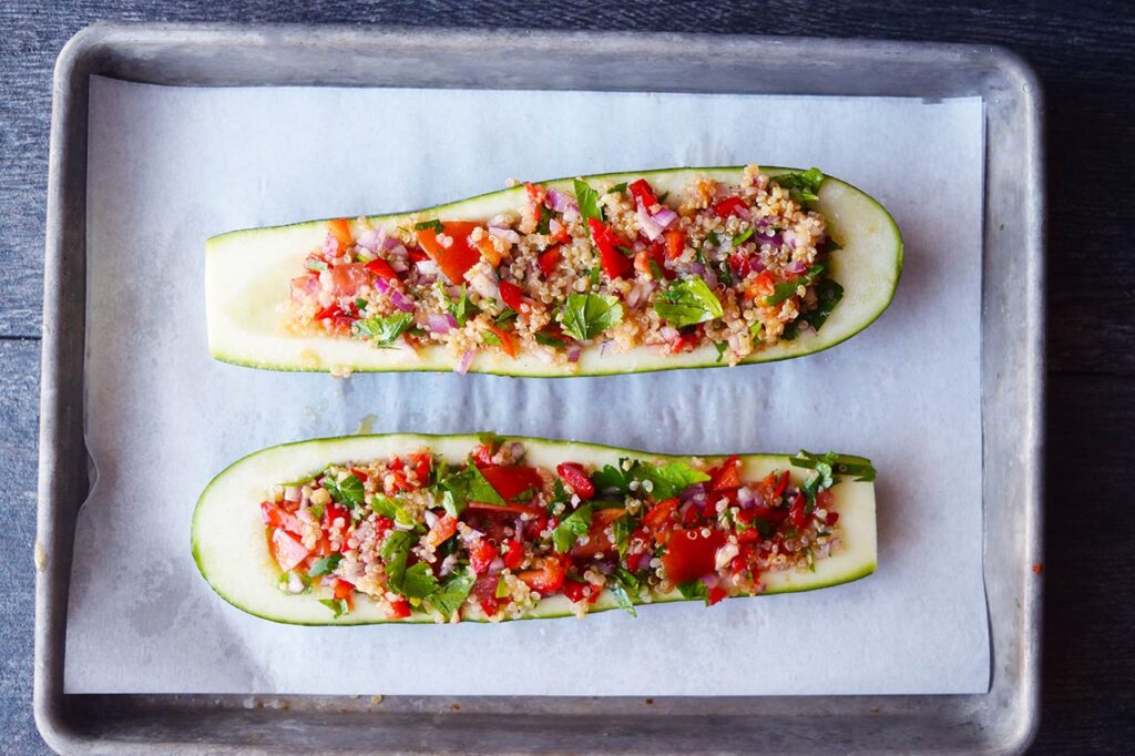 Two zucchini halves laying on a parchment-lined baking sheet, stuffed with quinoa stuffing.
