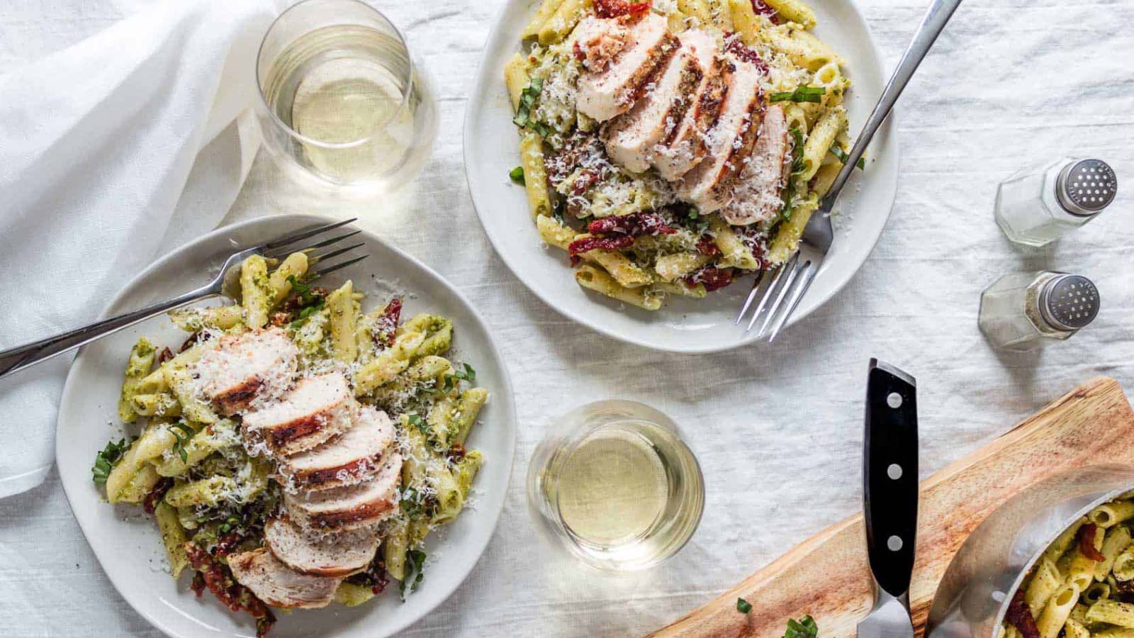 Two white plates with servings of creamy pesto pasta with chicken and sun dried tomatoes.